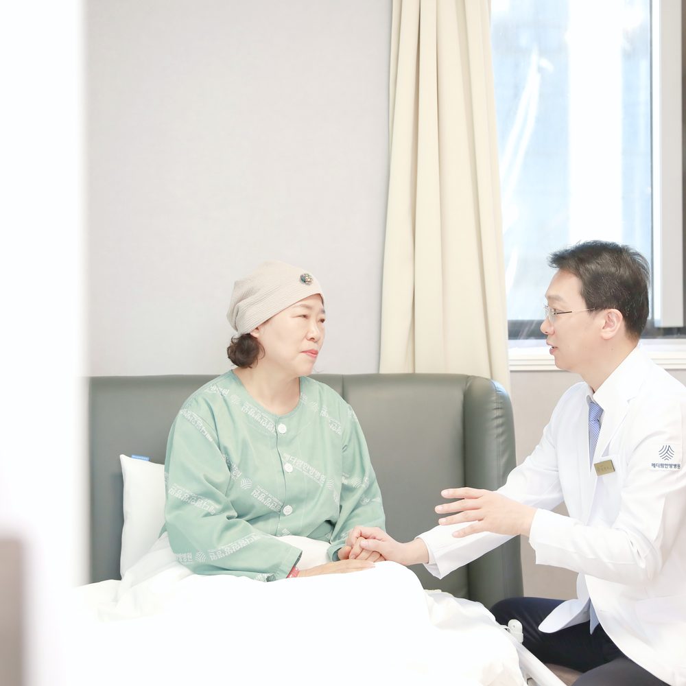 환자복을 입고 침상에 앉은 환자의 손을 잡고 원장이 요즘 피로와 식사 상태를 묻는 모습
