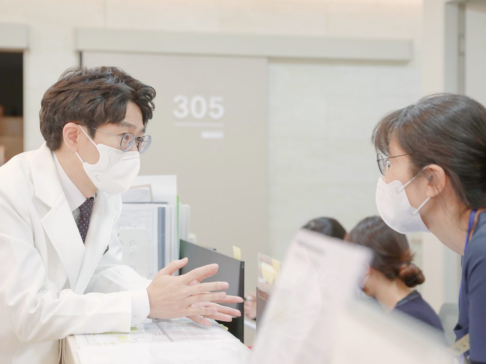 원장이 접수대 앞에서 환자복 차림의 환자와 마주 서서 다음 외래 검사 일정을 손짓으로 설명하는 모습