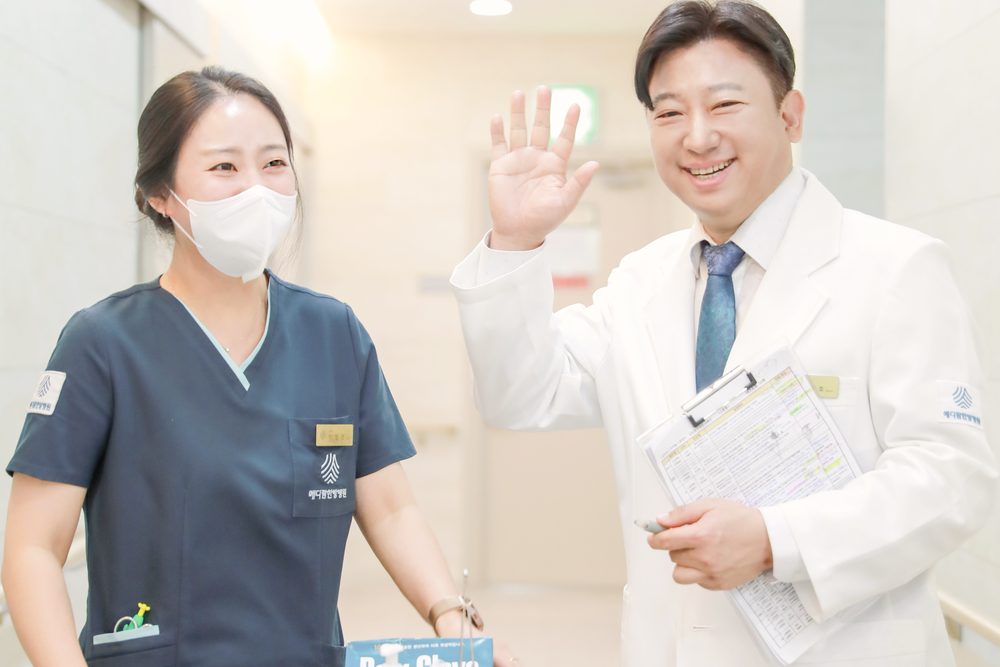병동 복도에서 원장이 간호사와 함께 환자에게 손을 들어 인사하며 오늘의 검사 일정표를 들고 있는 모습