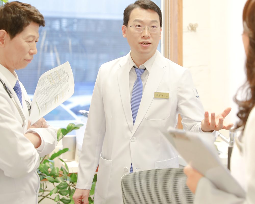 흰 가운을 입은 의료진 두 명이 병동 창가에서 진료 기록지를 들고 환자 상태를 함께 이야기하는 모습
