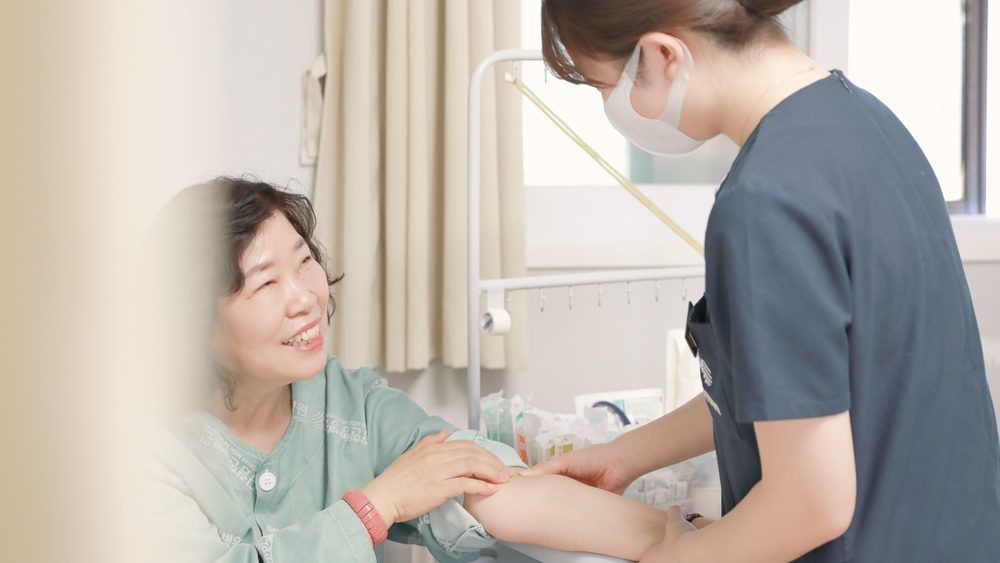 병실에서 간호사가 환자의 팔을 살피며 주사 부위를 확인하고, 환자가 웃으며 이야기하는 모습